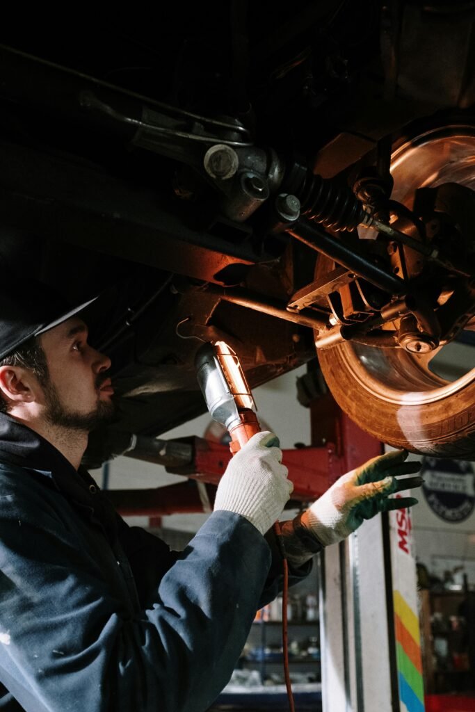 pexels-photo-4489737-4489737 Mechanic uses a flashlight to inspect car's suspension in a garage setting.